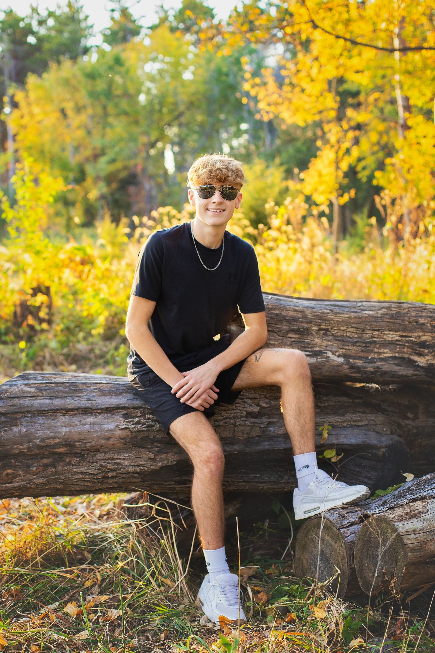 Smiling teen boy in black outfit and sunglasses sitting on a log surrounded by golden fall foliage.