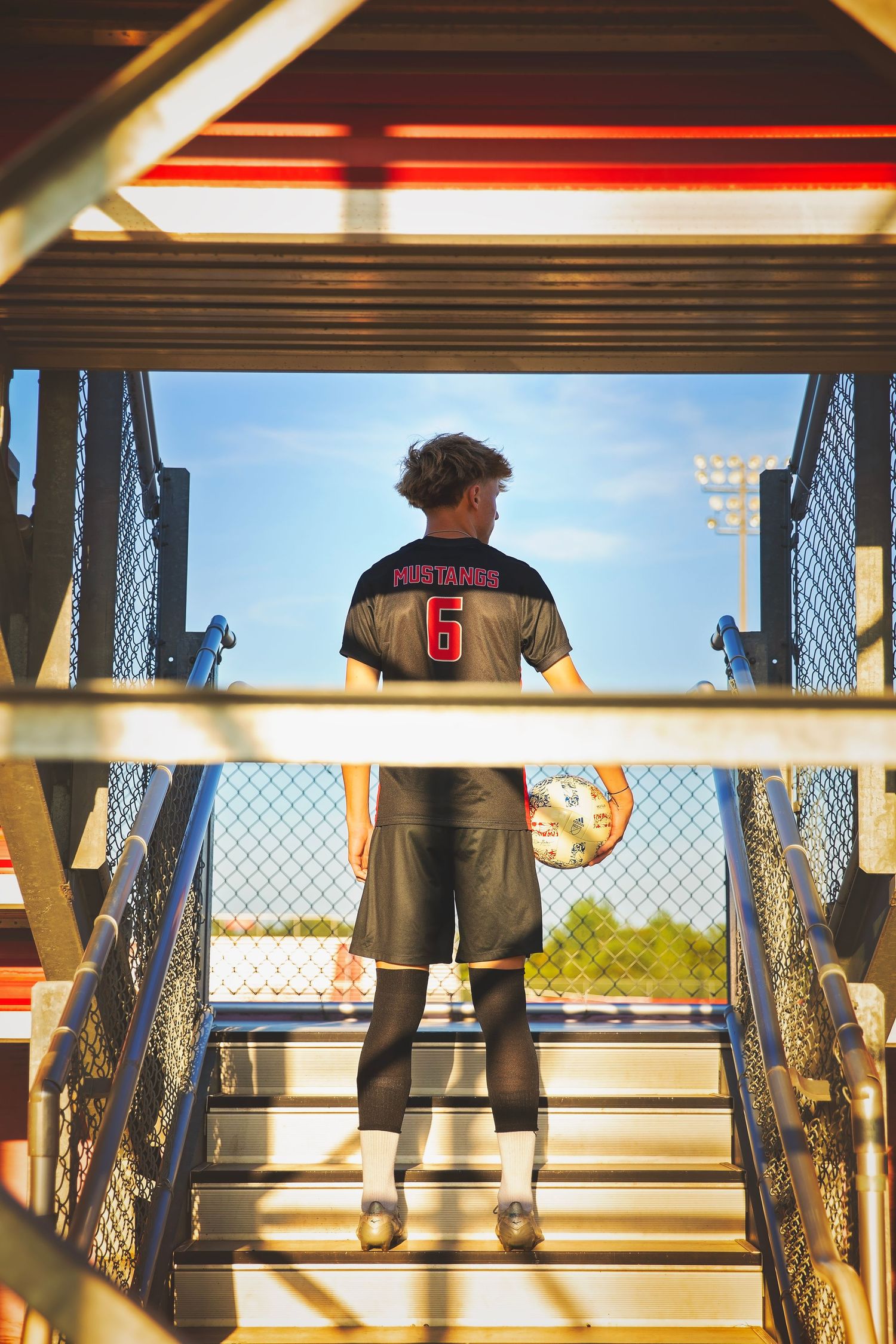 Soccer player wearing Mustangs number 6 jersey stands holding ball at top of stadium stairs.