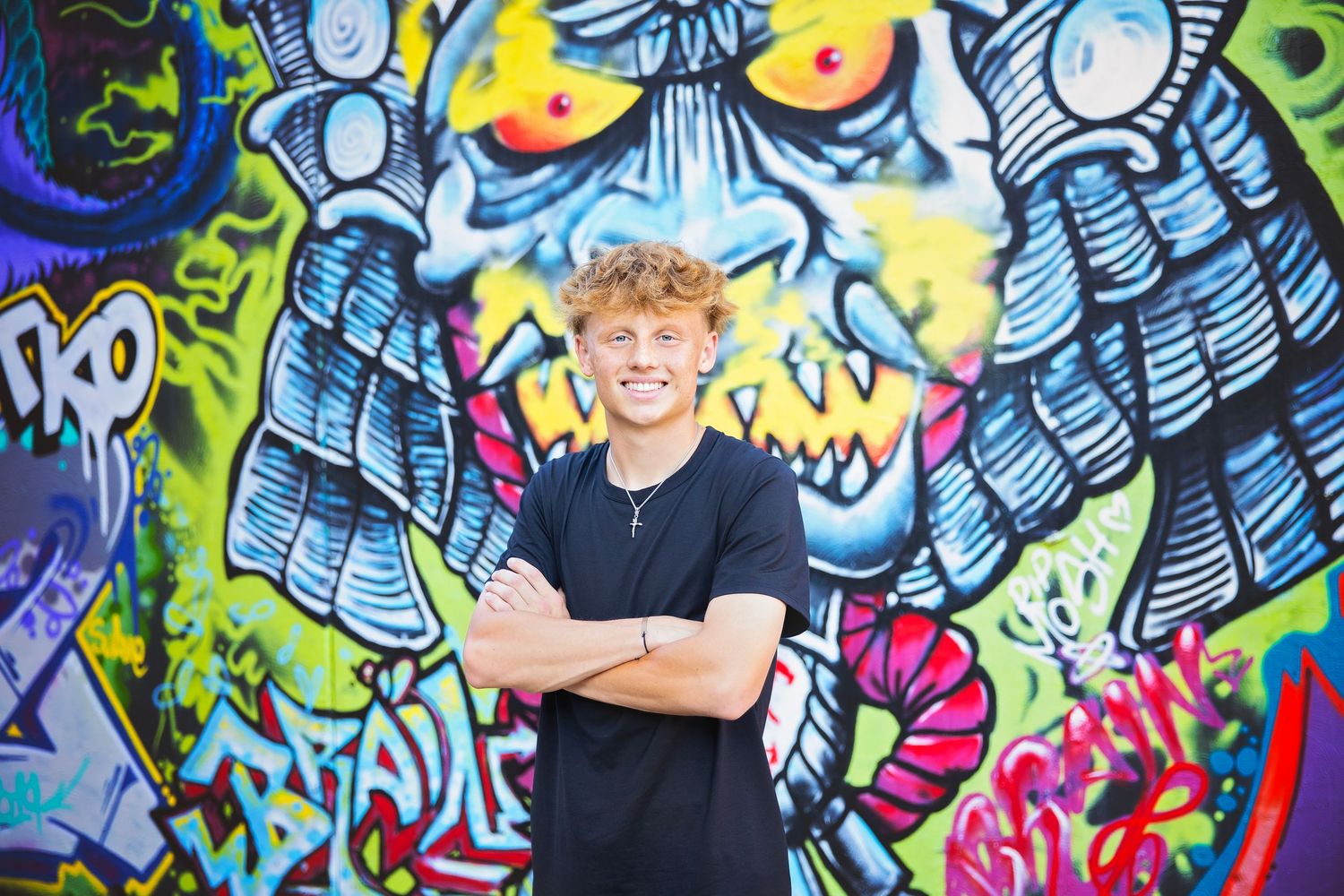 Smiling teen boy with curly hair stands arms crossed before vibrant colorful graffiti mural wall.
