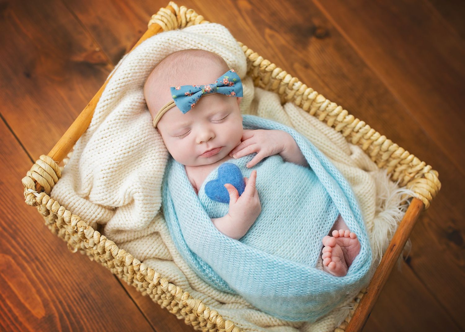 Sleeping newborn baby swaddled in blue knit wrap wearing bow headband lying in wicker basket.