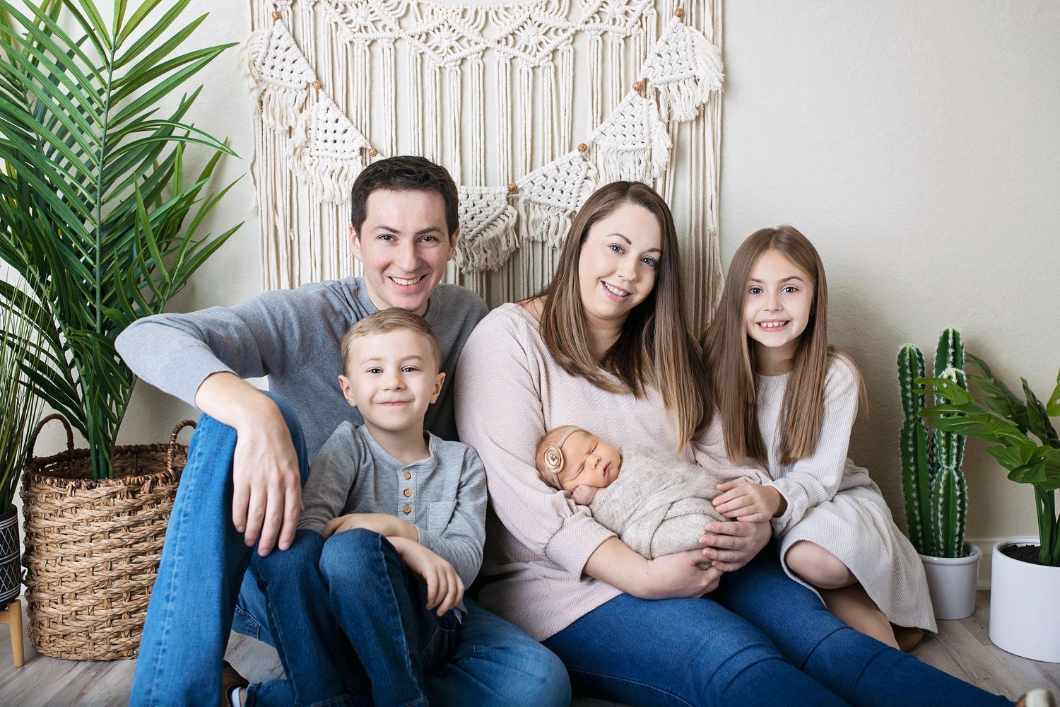 Family of four with two children posing together holding newborn baby in front of macrame wall decor.