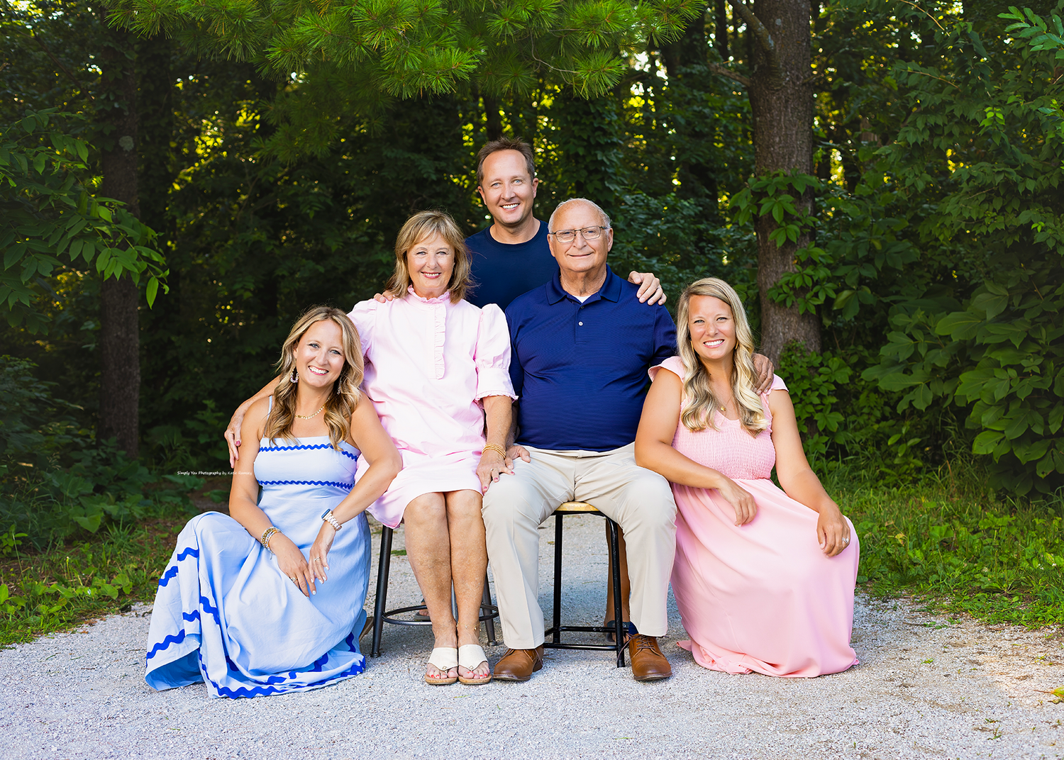 Family of five posing outdoors for a portrait, two women in pastel gowns seated, parents and son smiling among green trees.