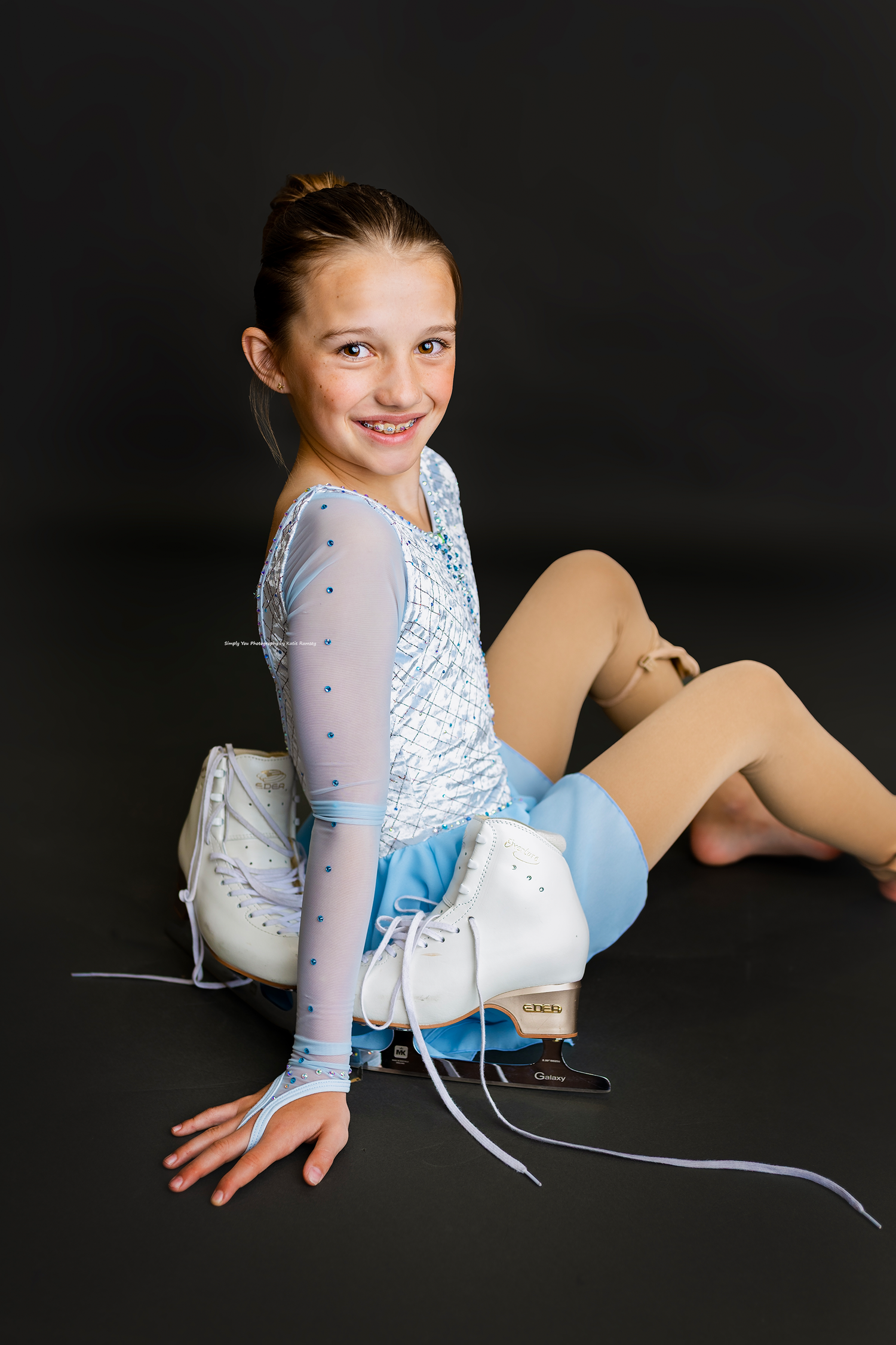Young figure skater in light blue dress and white ice skates poses smiling against dark background.