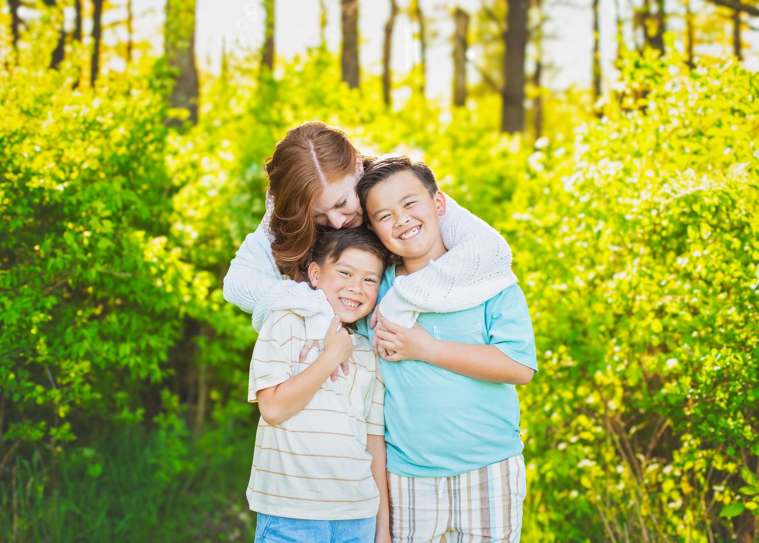 A woman hugs two laughing boys from behind in a sunny green outdoor setting.