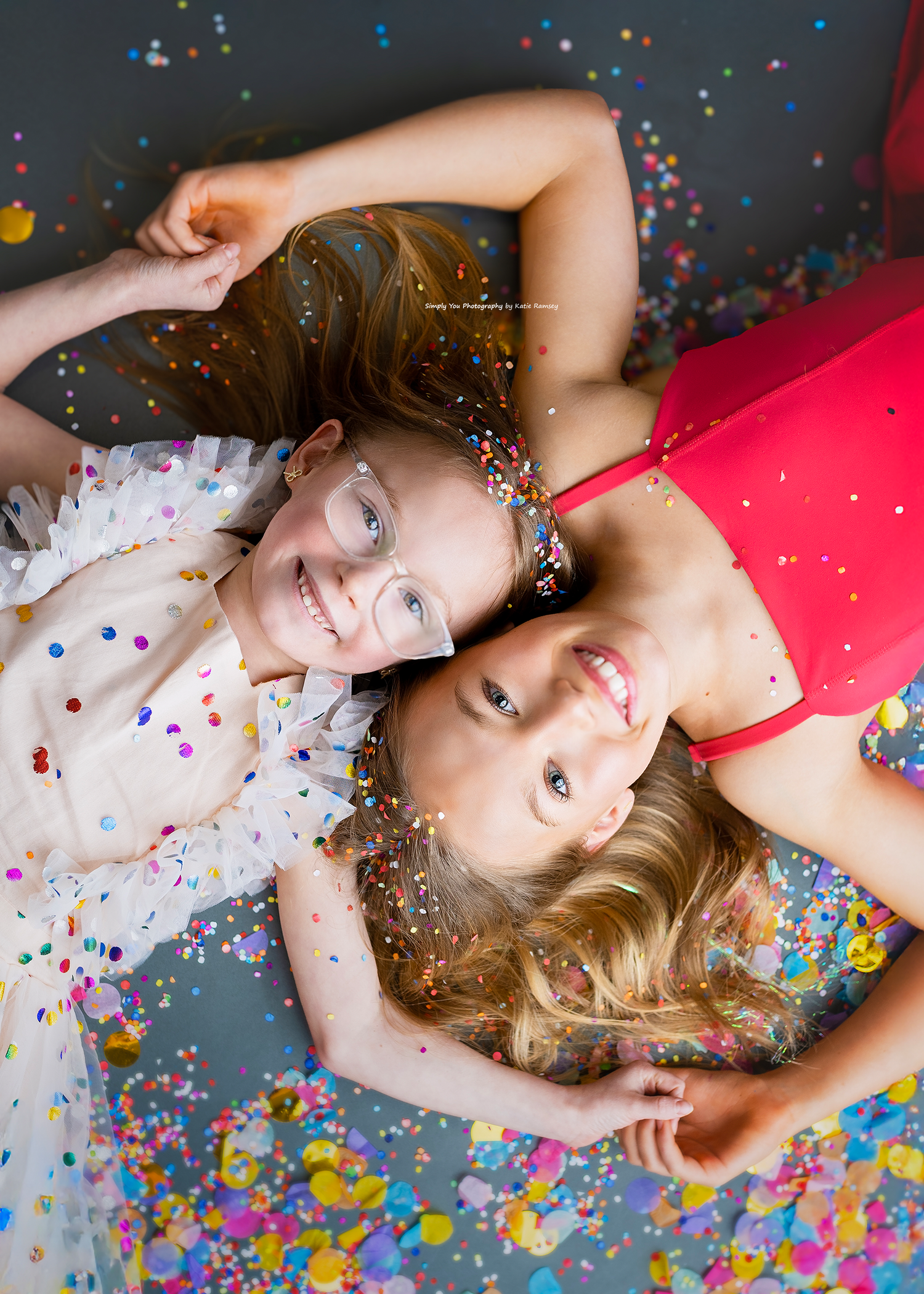 Two smiling girls lying on the floor surrounded by colorful confetti in a fun photography studio session.