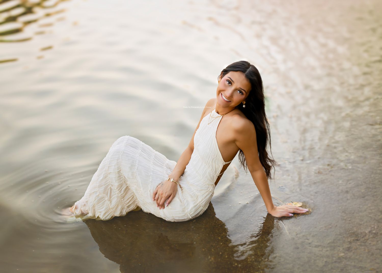 Young woman in white lace halter dress sitting gracefully in shallow lake water at golden hour.