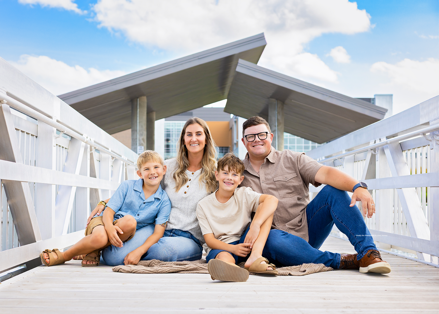 Happy family of five sitting on a white wooden bridge outdoors under a blue cloudy sky near a beach house.