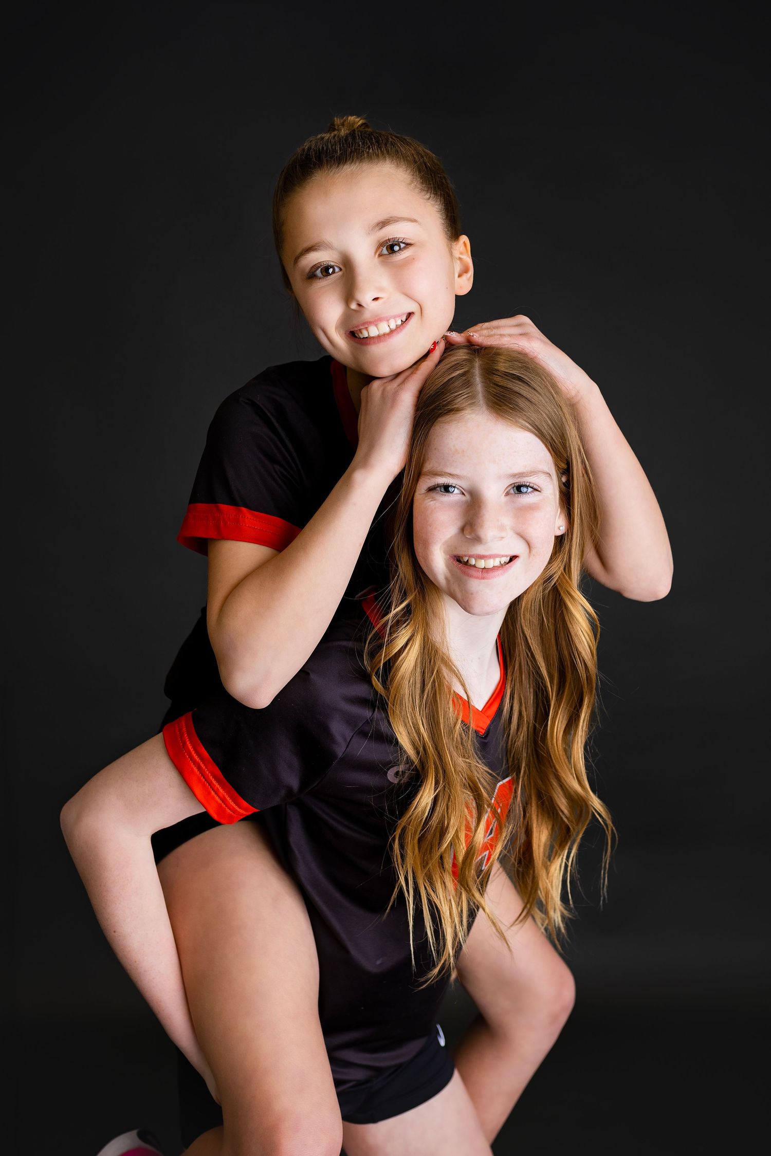 Two young girls in matching black and red volleyball uniforms smile together playfully in a dark studio setting.