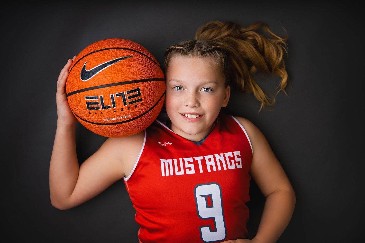Young girl in Mustangs number 9 jersey poses with a Nike Elite basketball on shoulder.