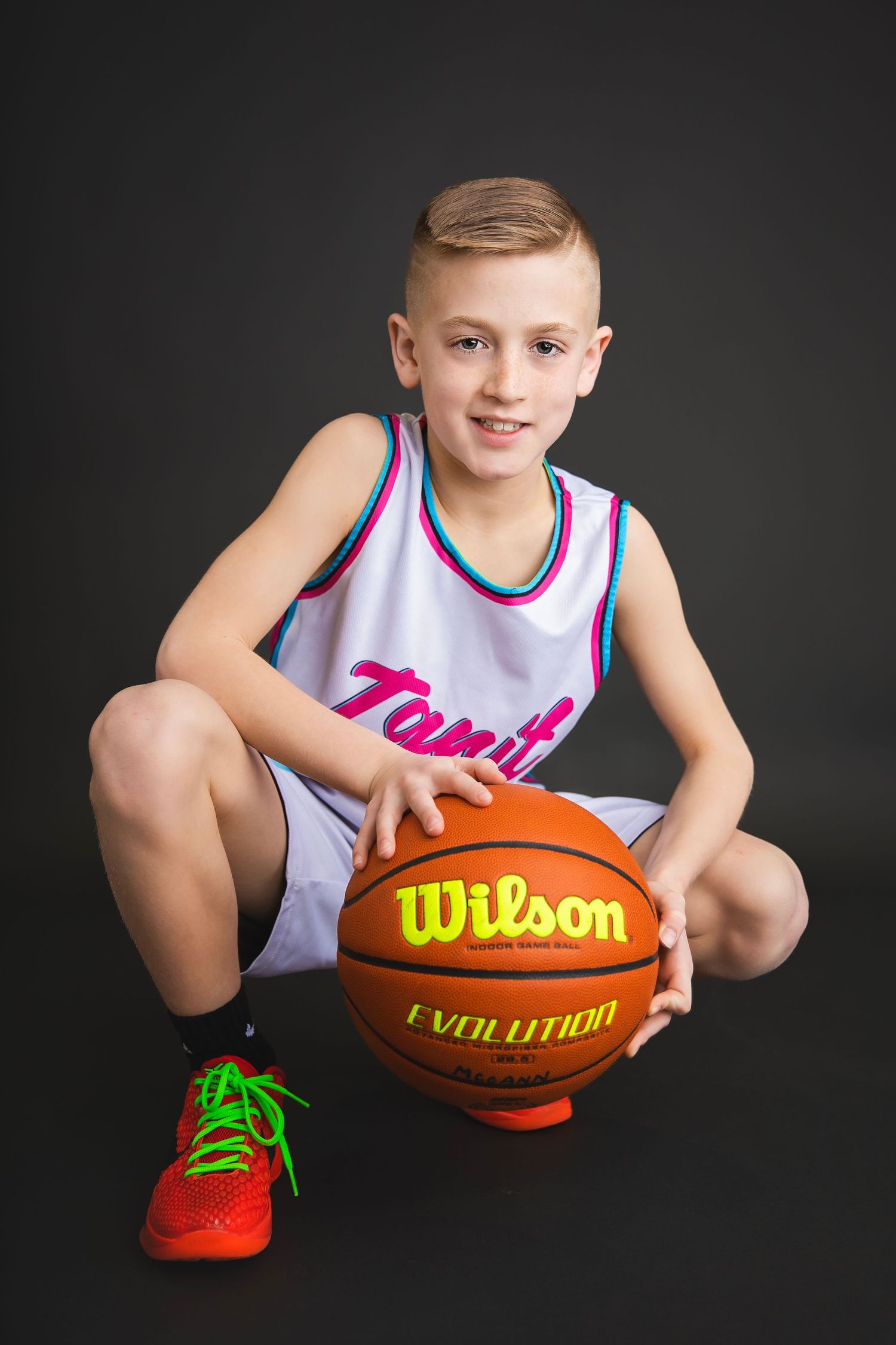 Smiling boy in Ignit uniform crouches holding Wilson Evolution basketball on floor.