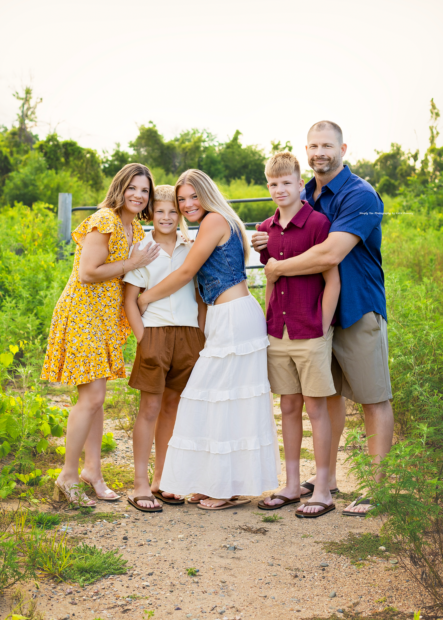 A happy family of five poses together on a sunny dirt path surrounded by lush green foliage.
