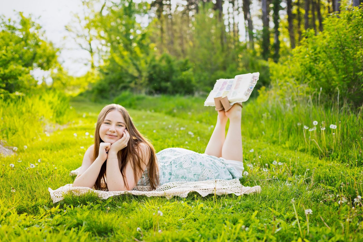 Young woman in floral dress lying on grass in sunny meadow reading a book outdoors.