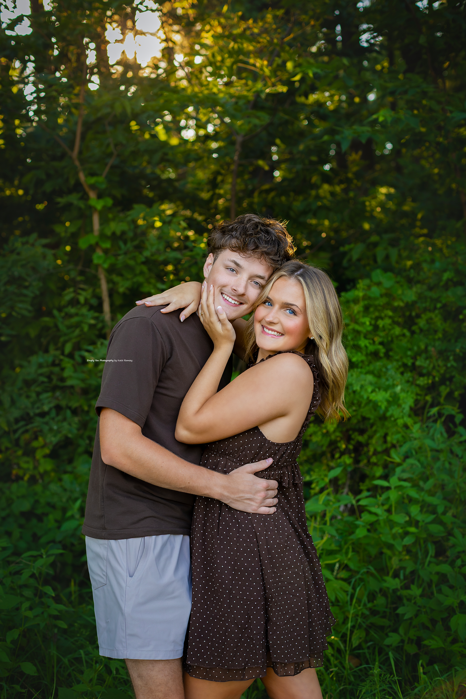 A smiling couple embraces outdoors, woman in brown dress hugs man in brown shirt, lush green trees in background.