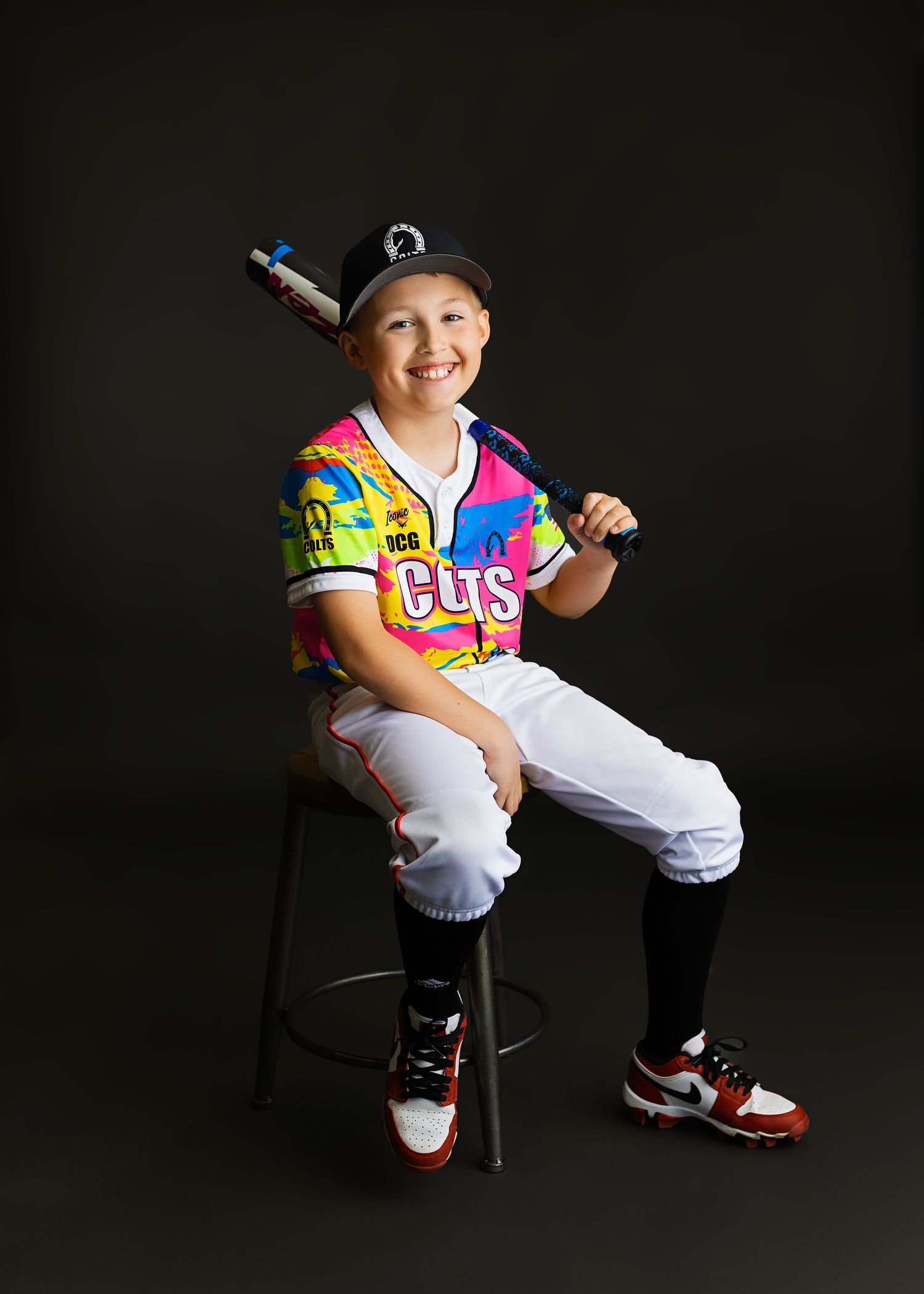 Young baseball player in colorful Cubs jersey and cap sits on stool holding bat for team photo.