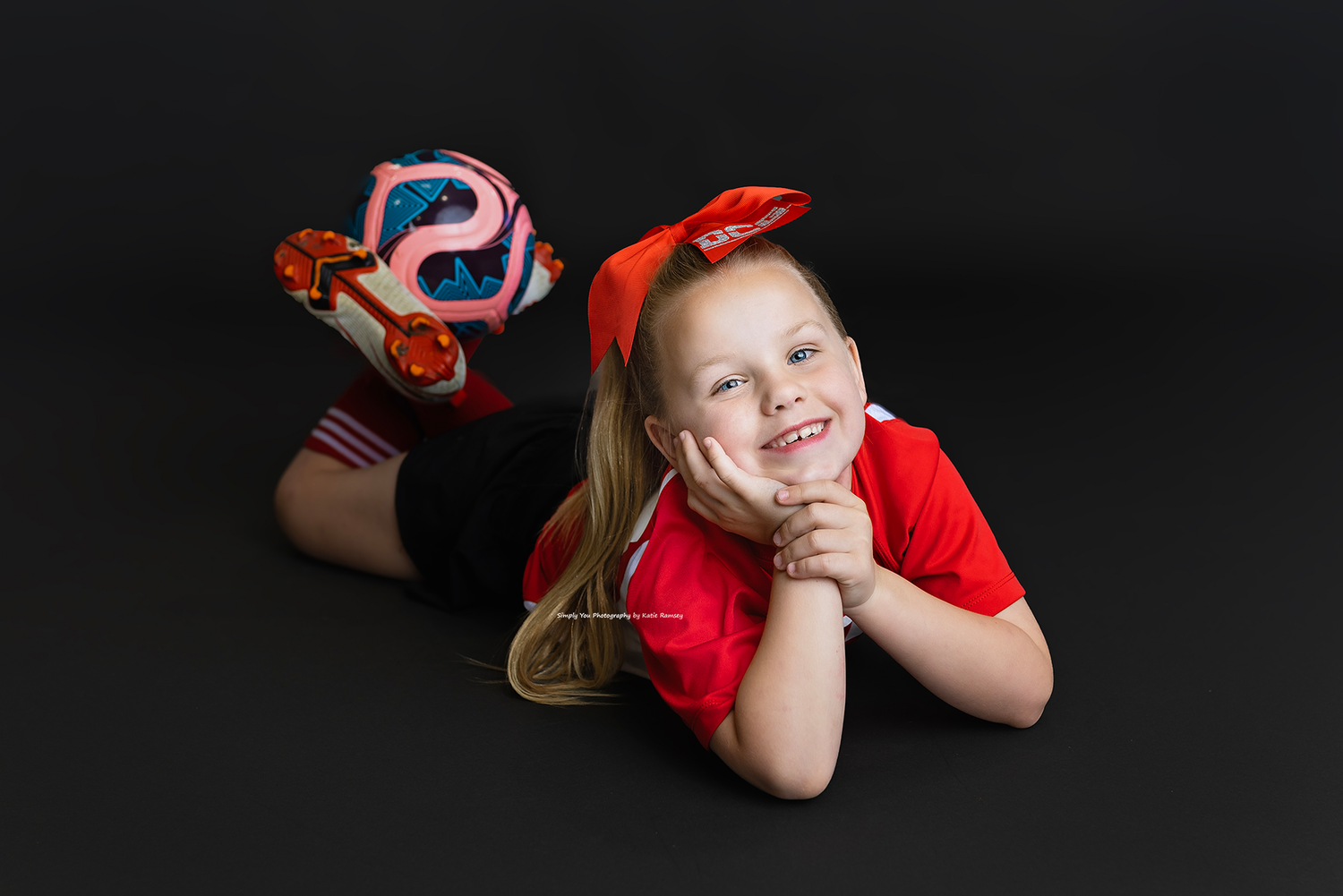 Smiling young girl in red outfit lying down wearing colorful roller skates on black background.