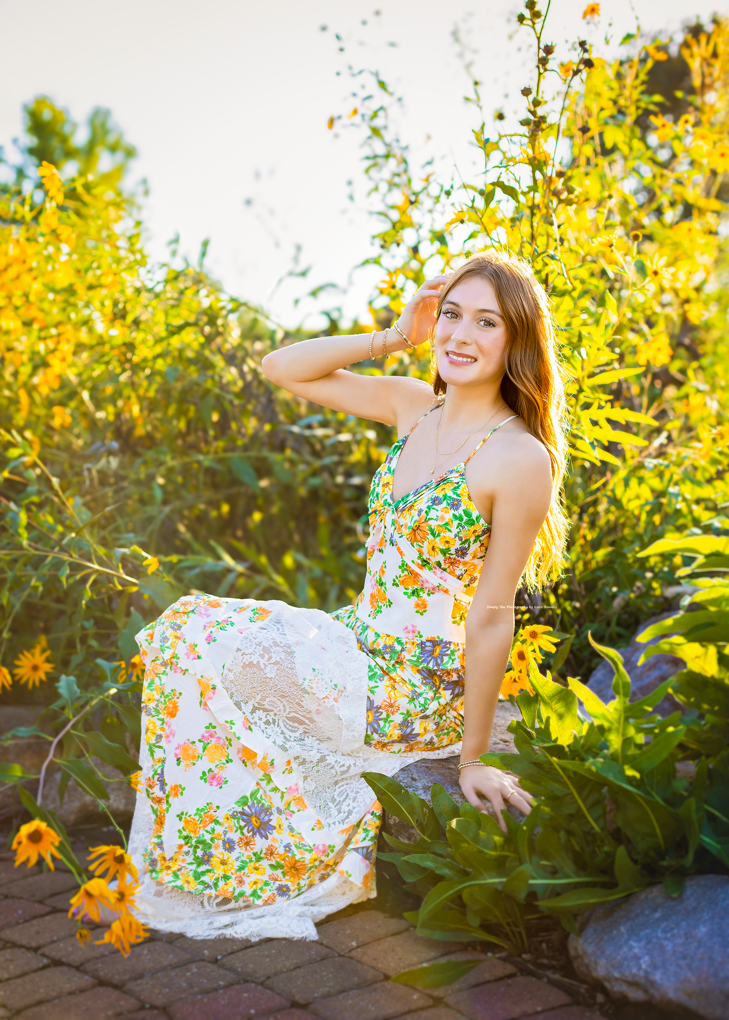 Young woman in floral maxi dress sitting among yellow wildflowers in a sunlit garden.