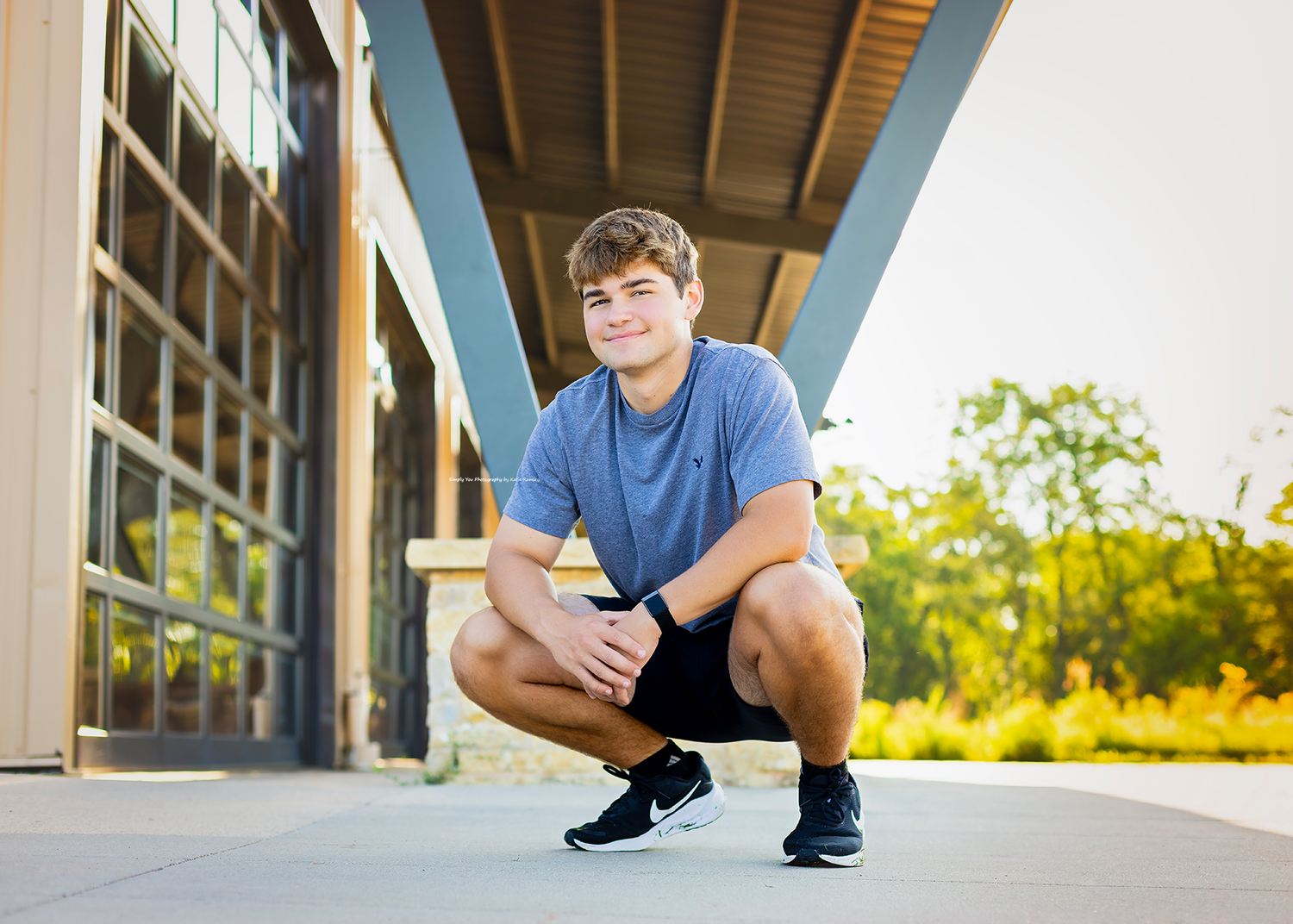 A smiling teenage boy crouches confidently outdoors in a blue shirt and black shorts for senior portrait photos.