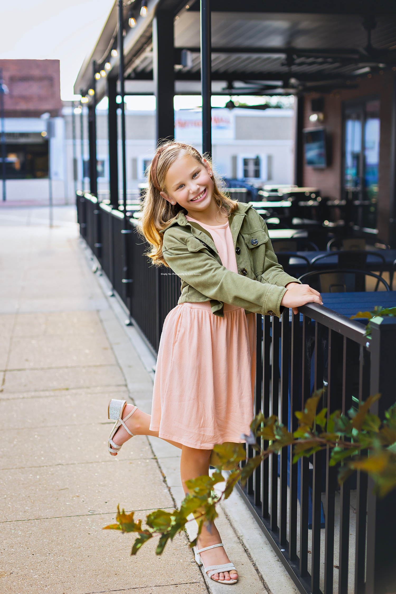 Smiling girl in pink dress and green jacket leaning on black railing outside a restaurant patio.