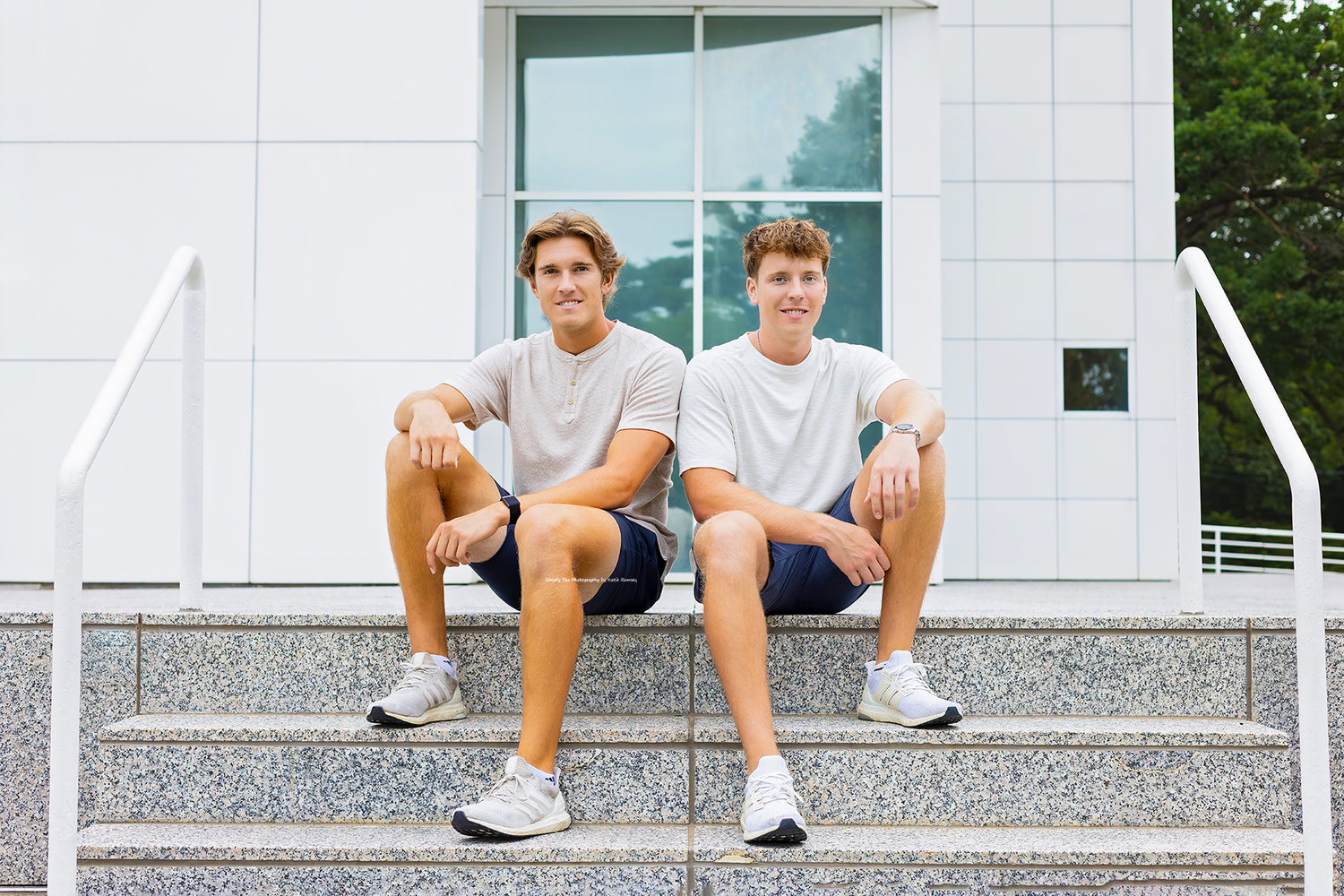 Two young men in matching white t-shirts and navy shorts sit together on outdoor steps smiling.