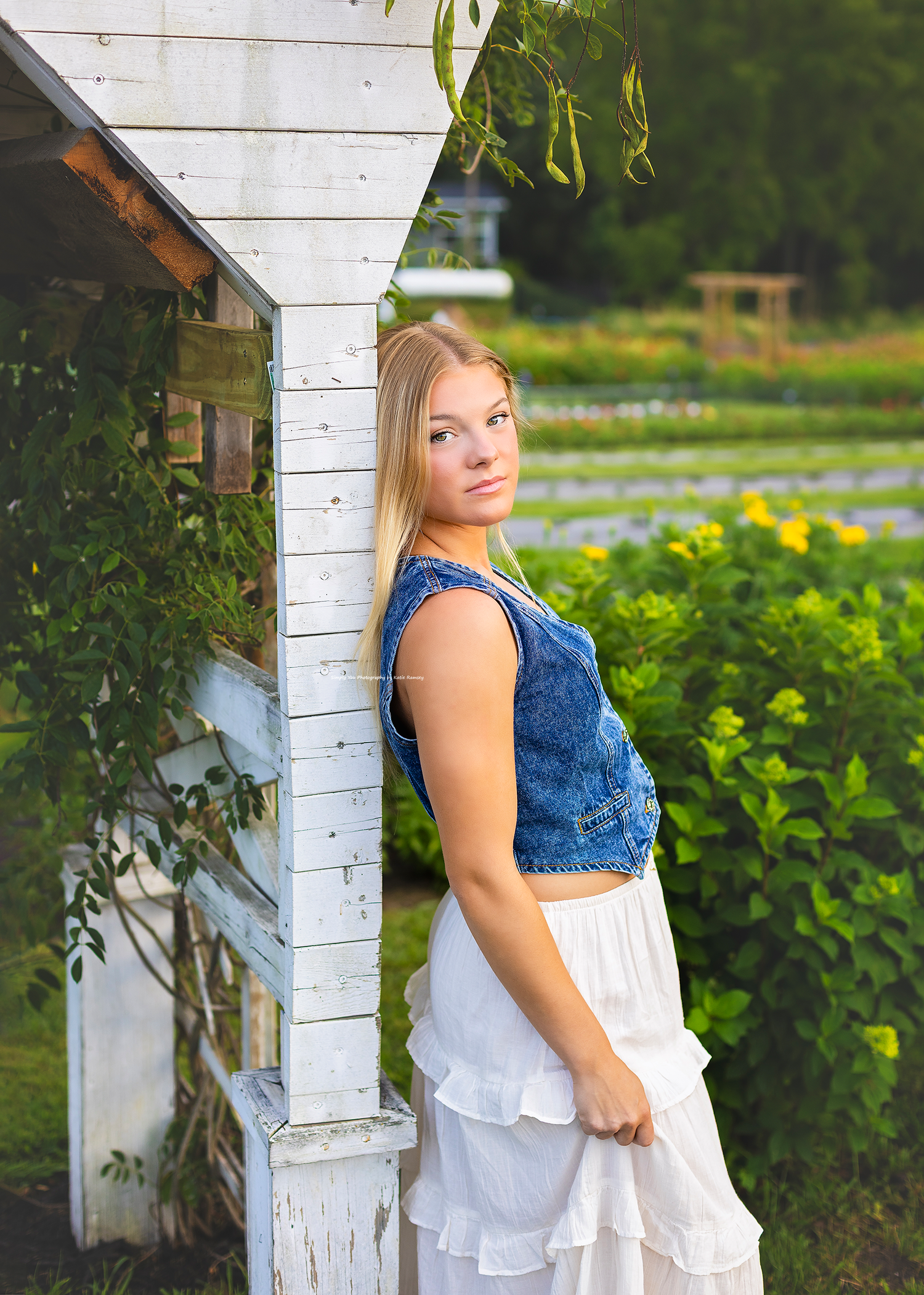 Young blonde woman in denim vest and white skirt posing by rustic white post with garden background.