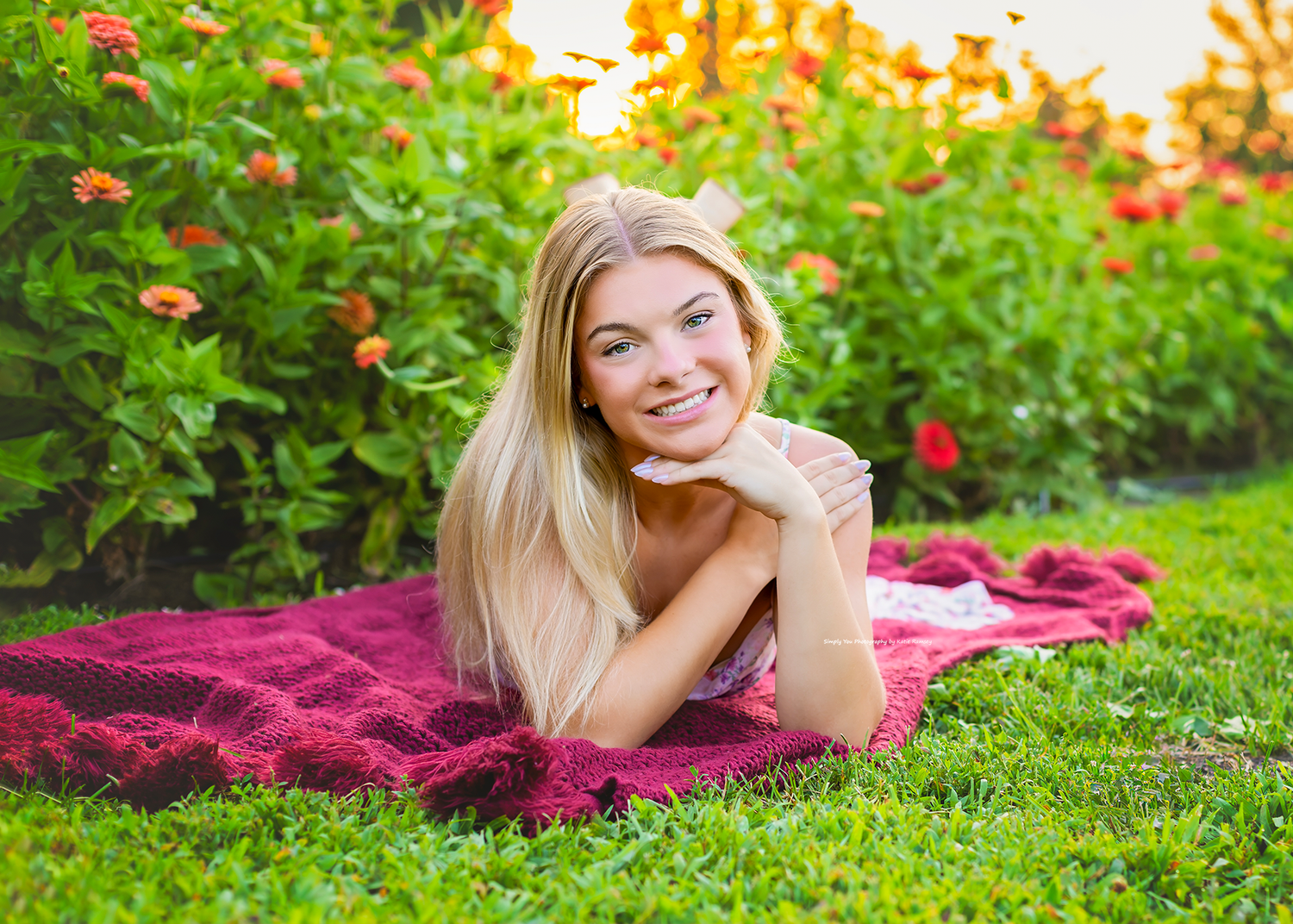 Smiling blonde woman lying on a red blanket in a lush green garden with colorful flowers in bloom.