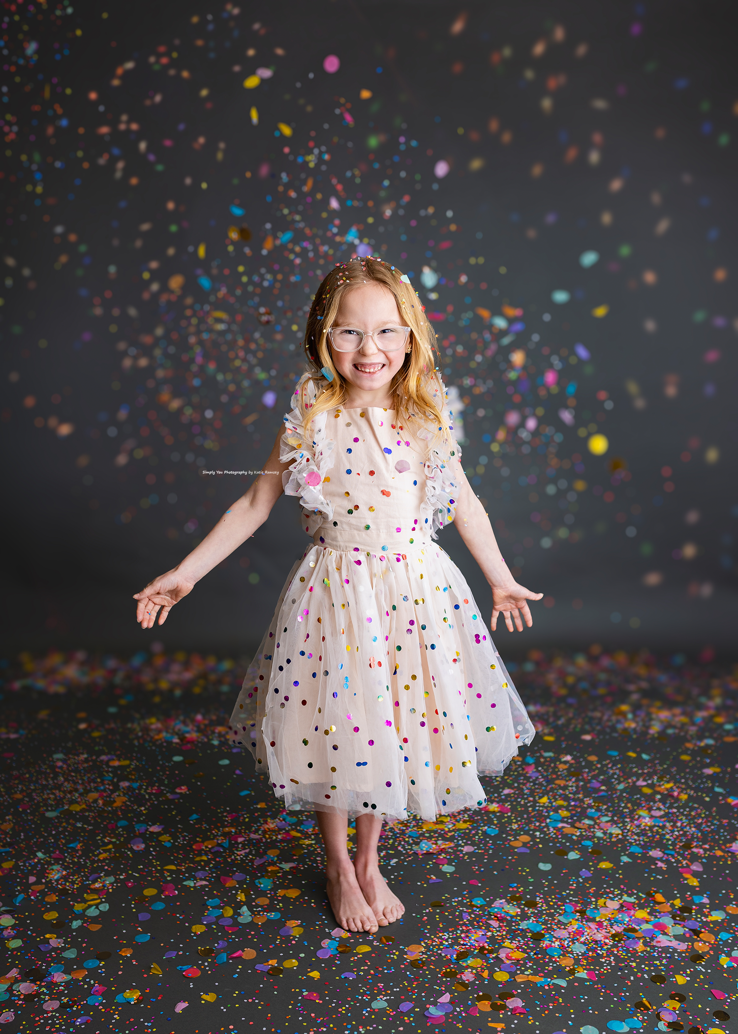 Happy redheaded girl in polka dot tulle dress stands barefoot dancing in falling rainbow confetti.