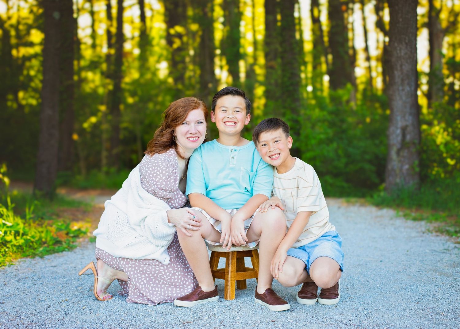 A smiling woman crouches beside two boys on a gravel path surrounded by tall green trees.