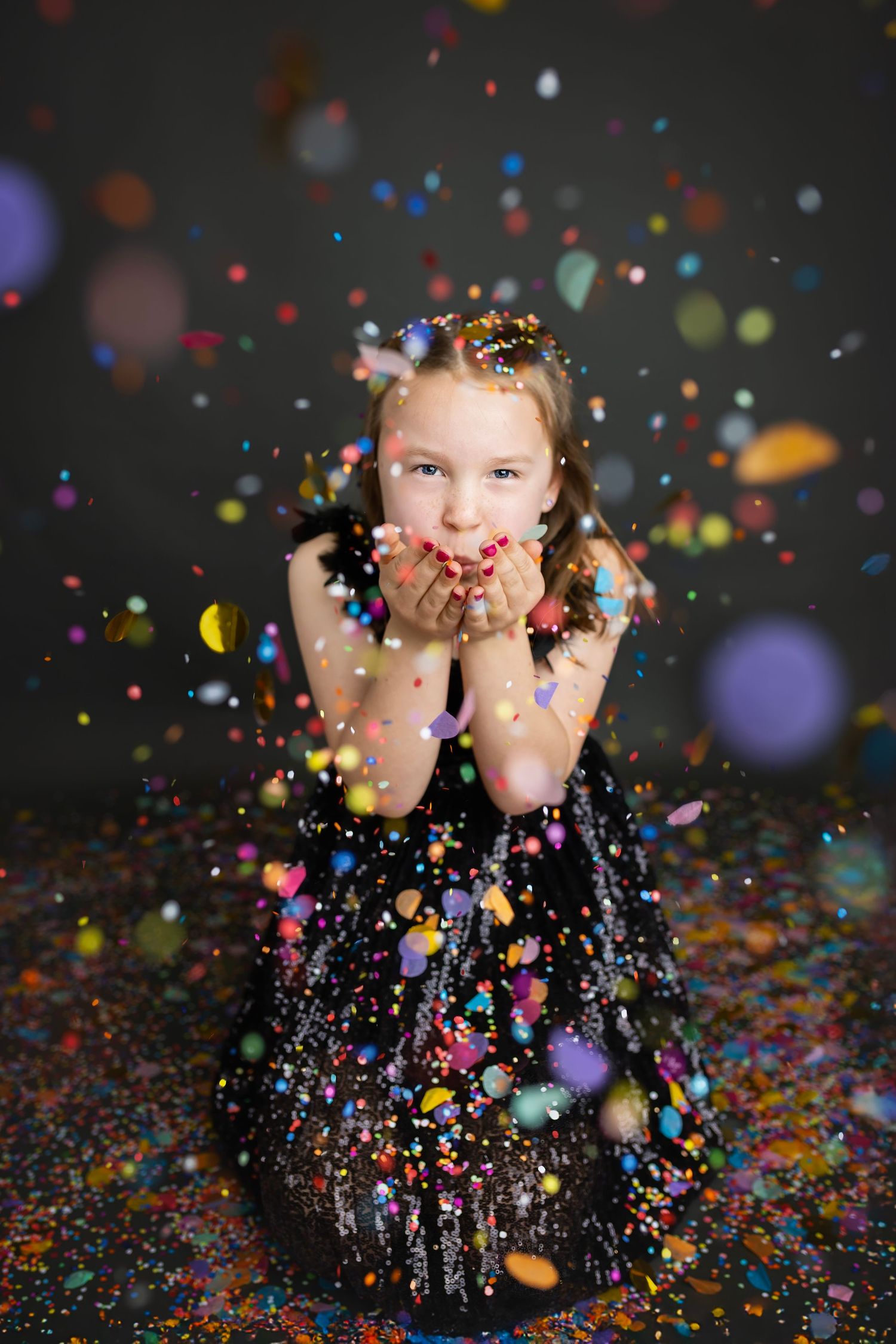 Young girl in black sequin dress blowing colorful confetti from her hands in a studio setting.