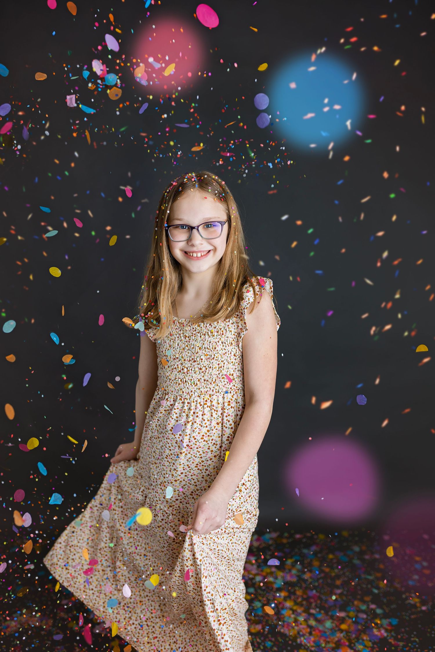 Smiling girl in floral sundress twirling among falling colorful confetti in a dark studio.