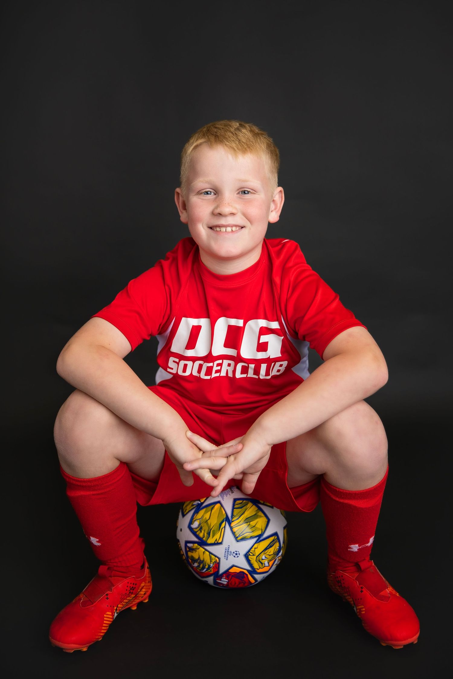 Smiling boy in red DCG Soccer Club uniform sitting on a soccer ball against a dark background.