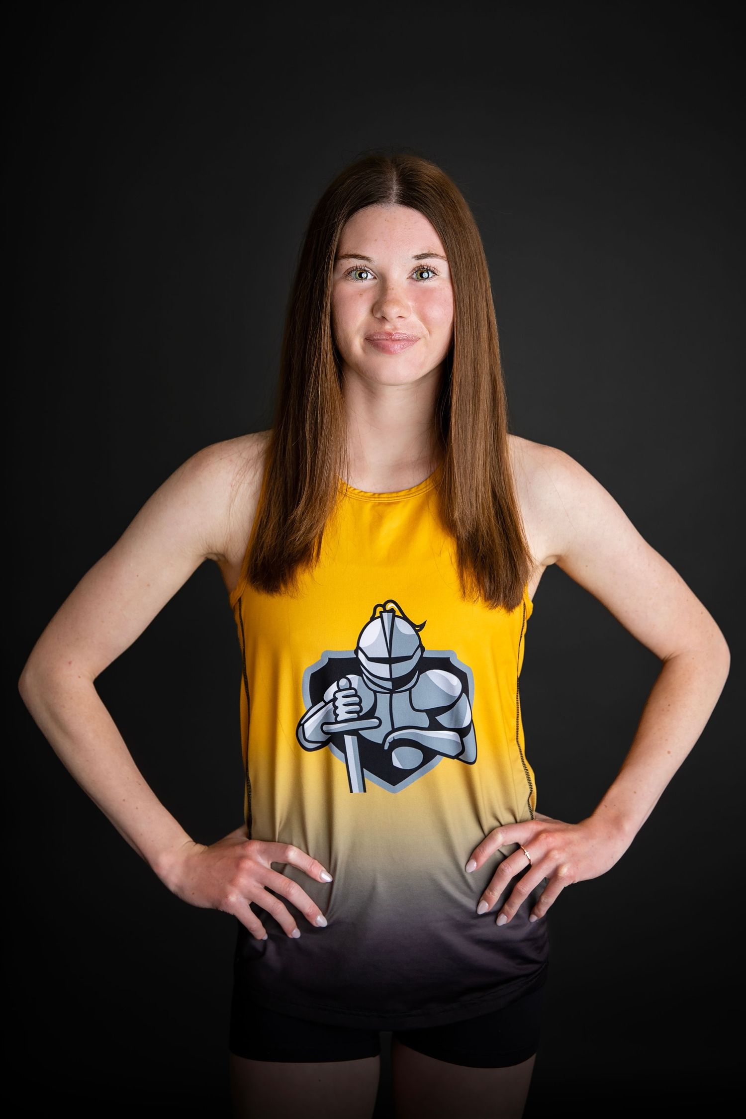Female athlete in gold knight-logo tank top poses with hands on hips against dark background.