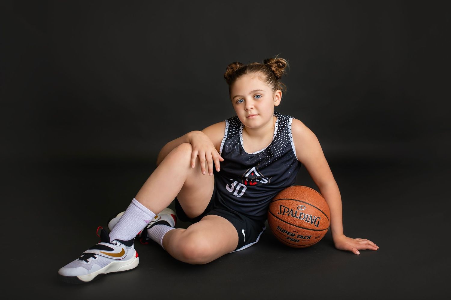 Young girl in navy basketball uniform number 30 sitting beside a Spalding basketball on dark background.