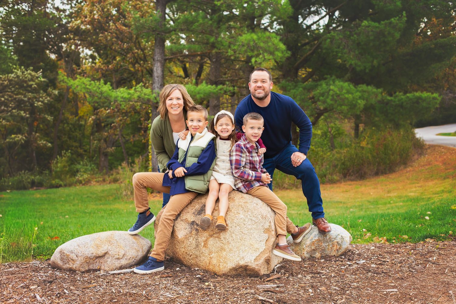 A smiling family of five poses together on large rocks in a lush green park setting.