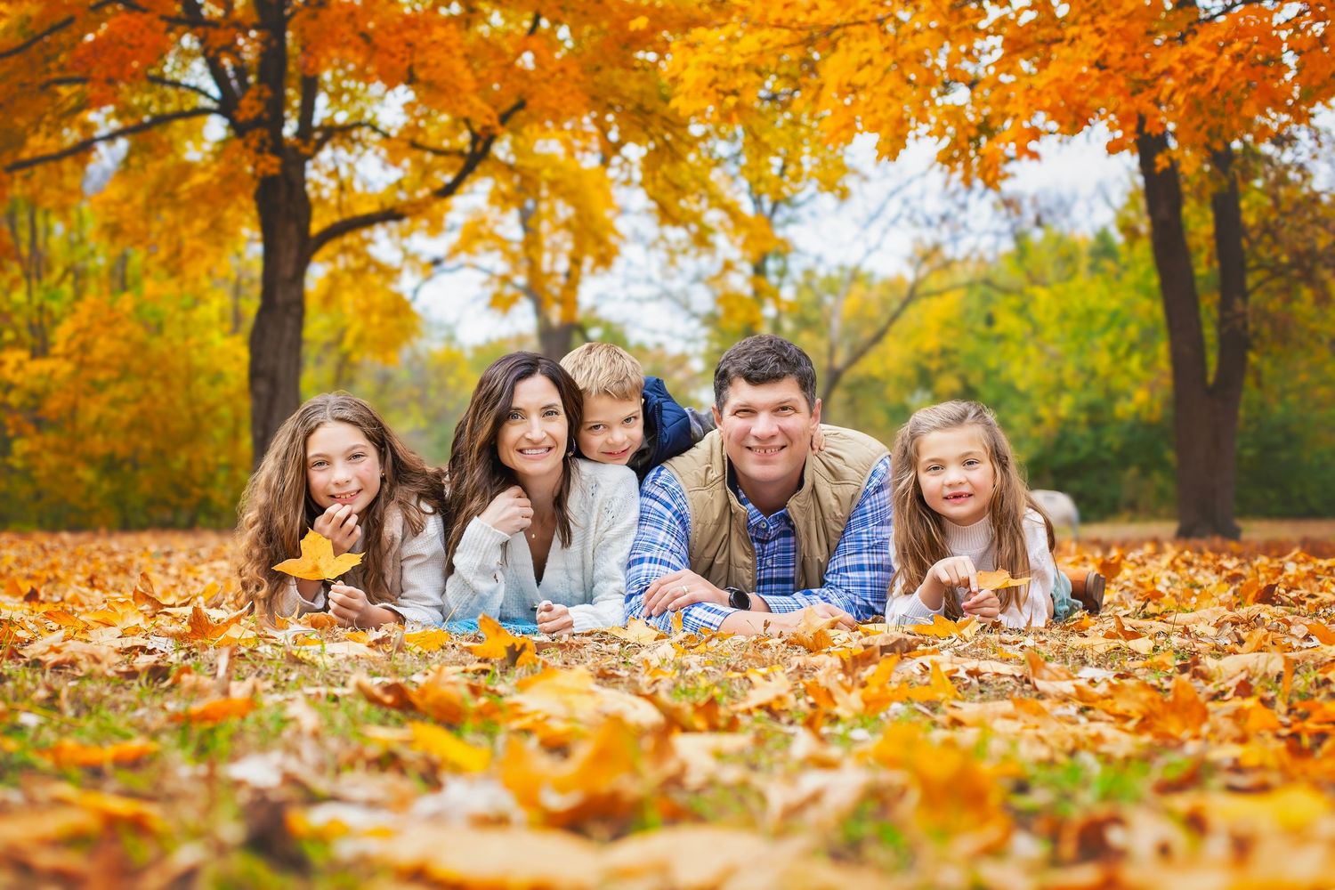 Happy family of five lying in autumn leaves under colorful fall trees.