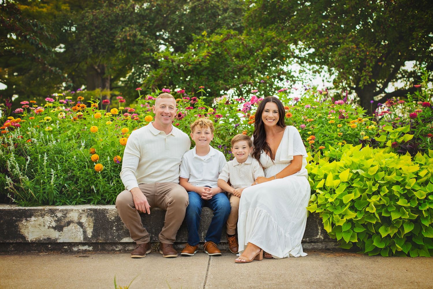 Happy family of four sitting together in front of a lush colorful flower garden outdoors.