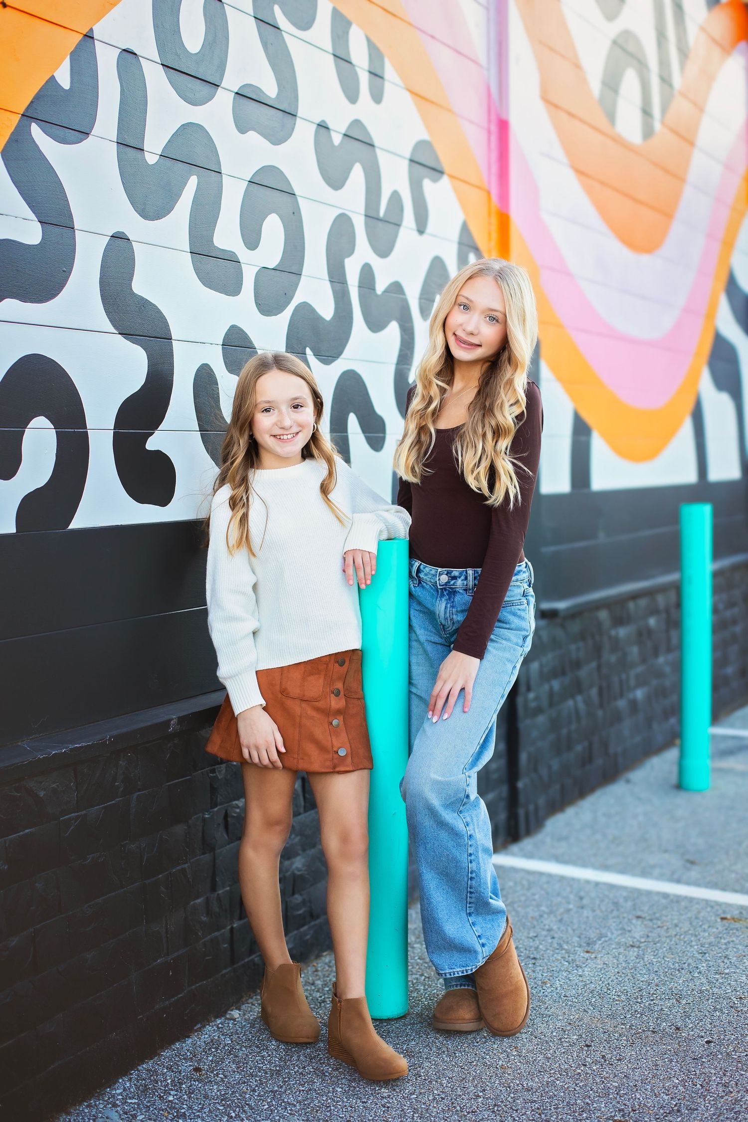 Two smiling sisters pose together in front of a colorful abstract mural on an urban street.