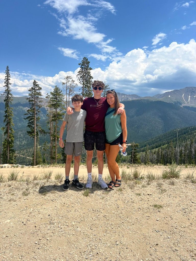 Three people smiling together on a mountain trail with scenic forest and peaks in background.