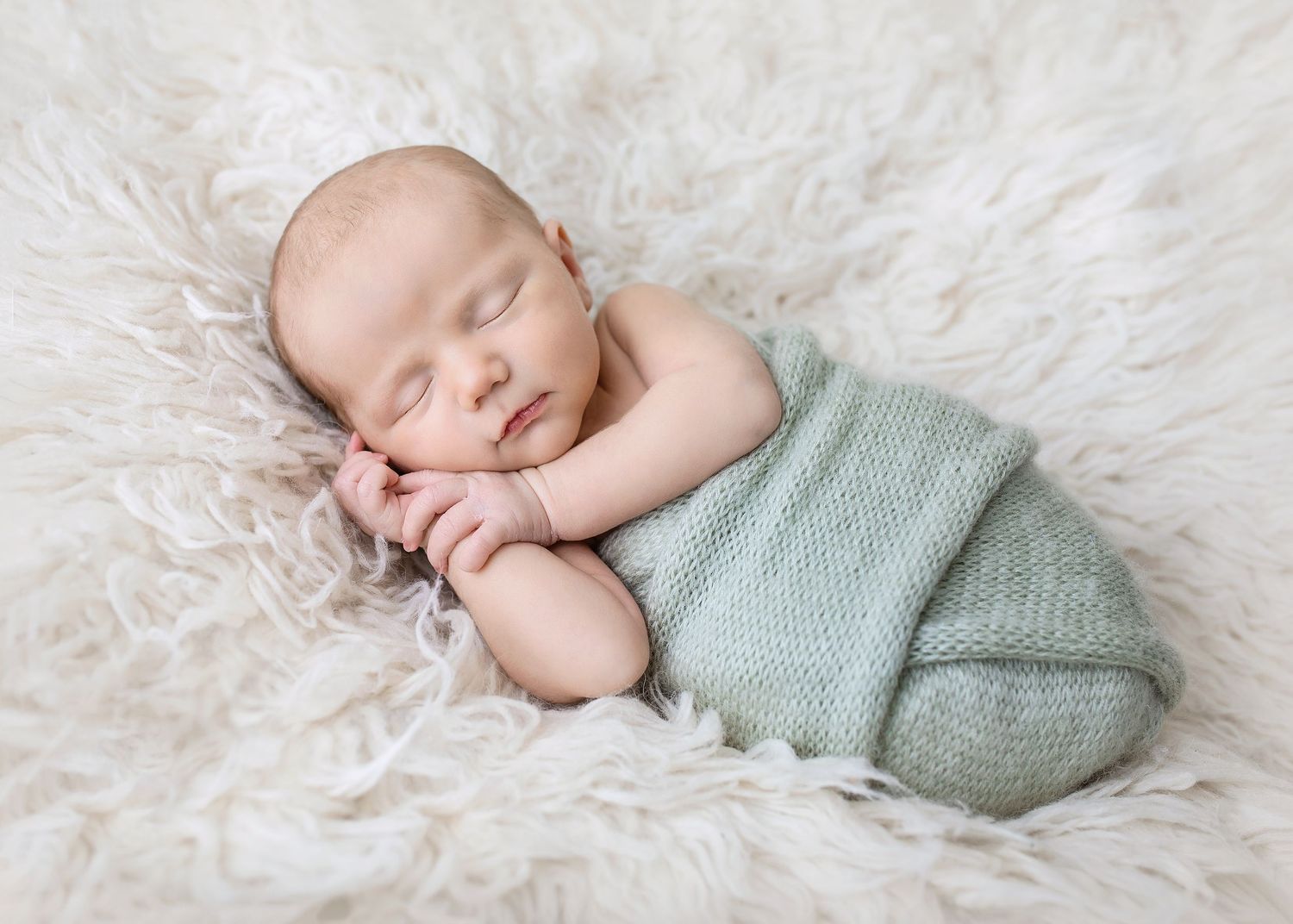 Sleeping newborn baby swaddled in a soft mint green knit wrap on a fluffy white fur blanket.