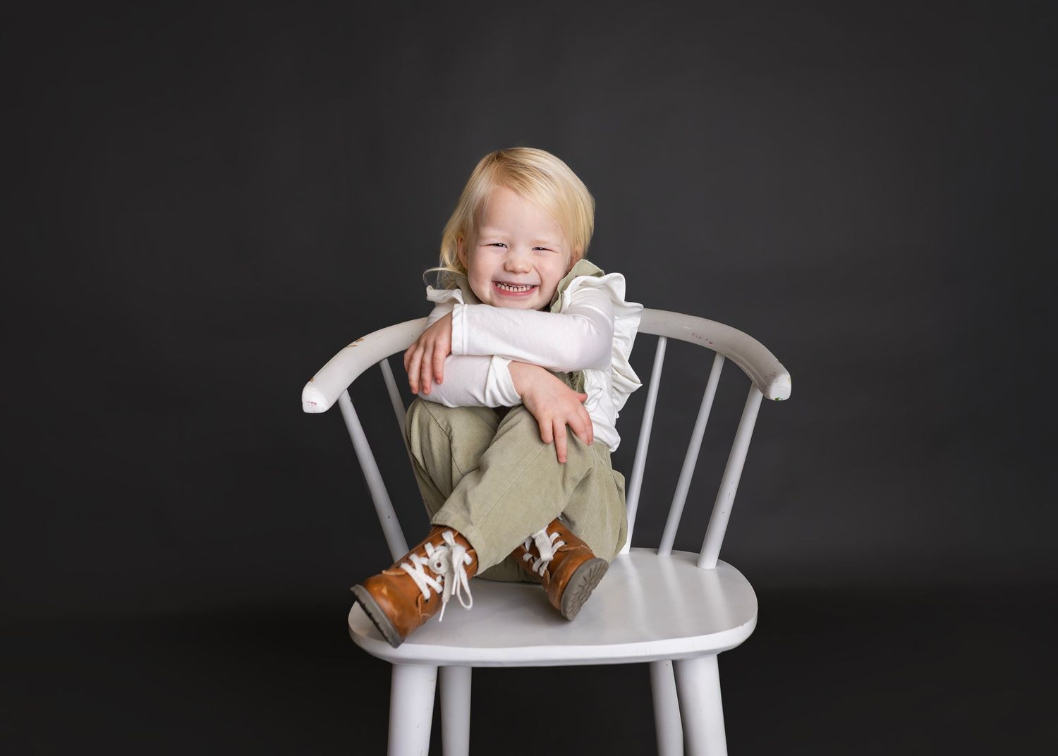 Cute toddler in green overalls sitting on white wooden chair laughing in studio portrait photo.