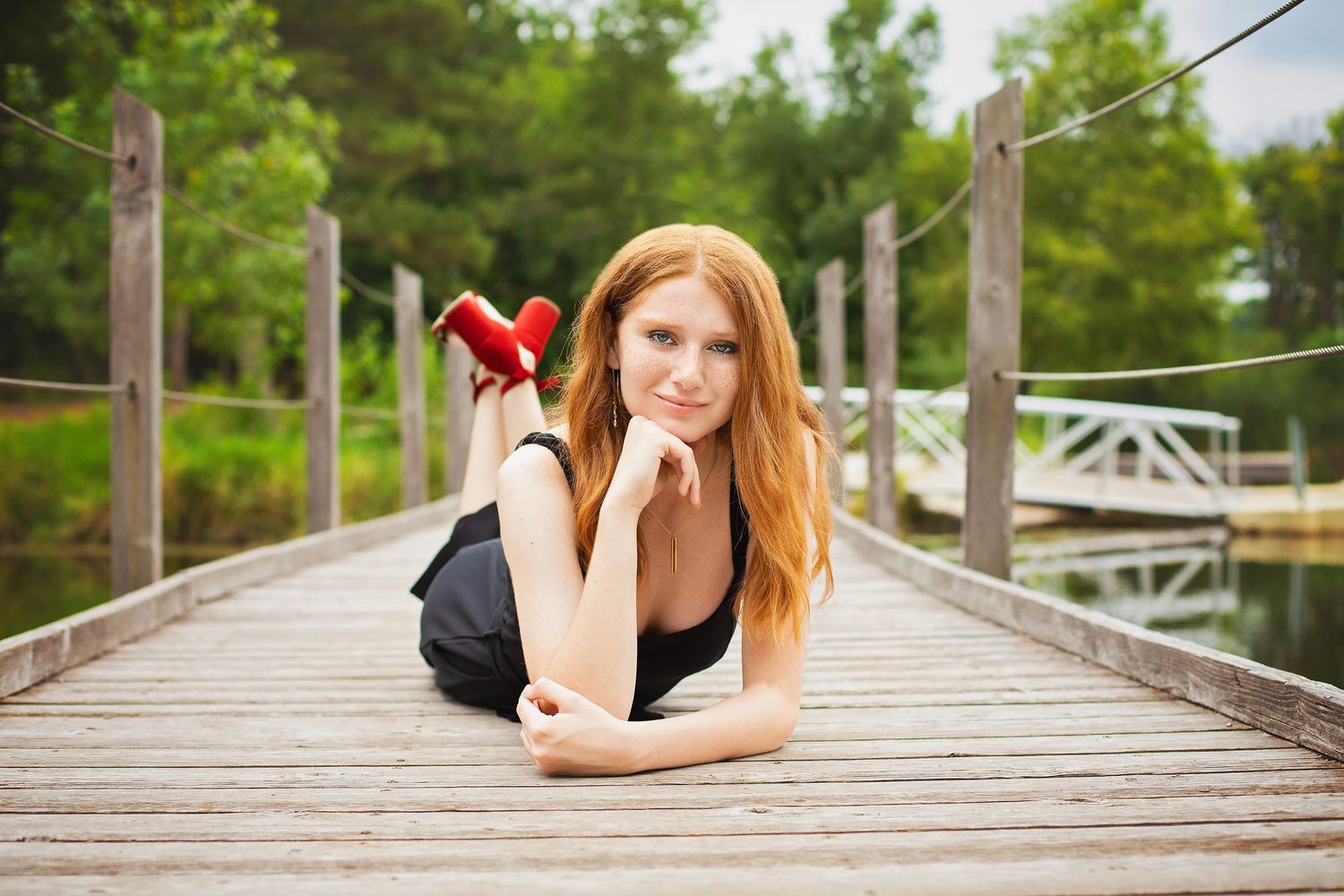 Red-haired teen girl lies on a wooden dock holding a red object near a calm lake.