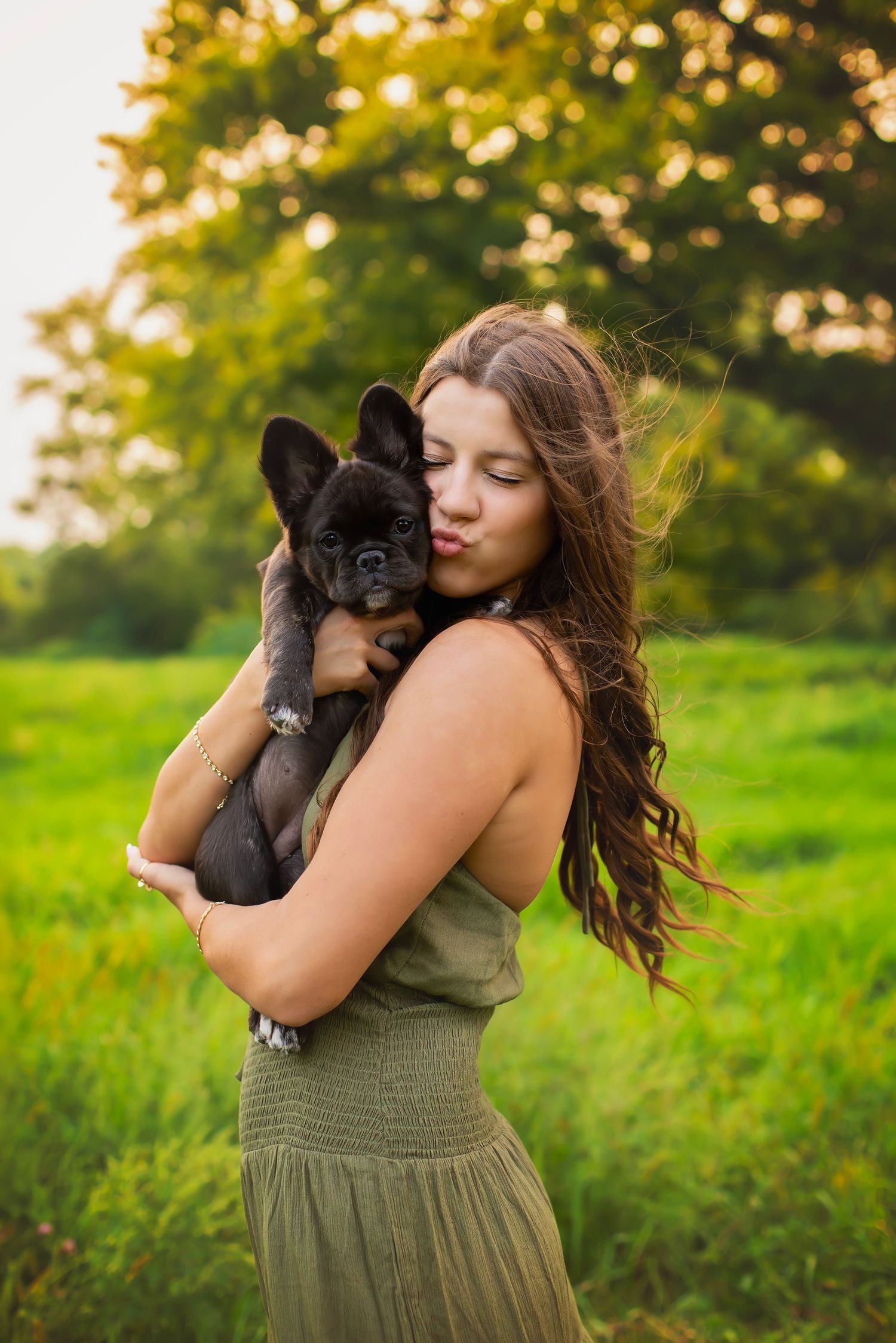 Young woman in olive dress holds black French bulldog puppy outdoors in green field at golden hour.