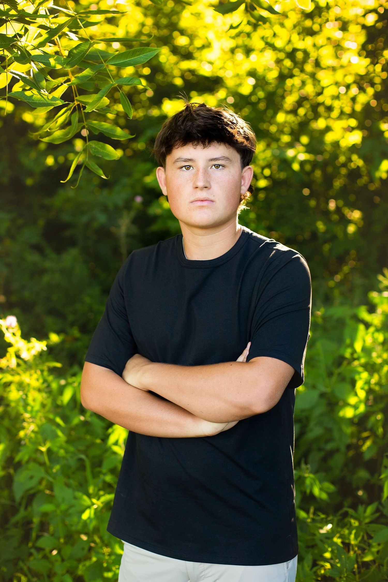 Teenage boy with arms crossed standing confidently in front of bright green sunlit foliage.