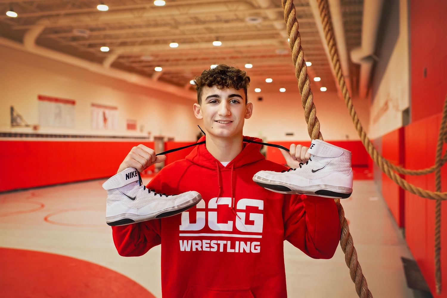 Teen in red DCG Wrestling hoodie holding up white wrestling shoes inside a gymnasium.