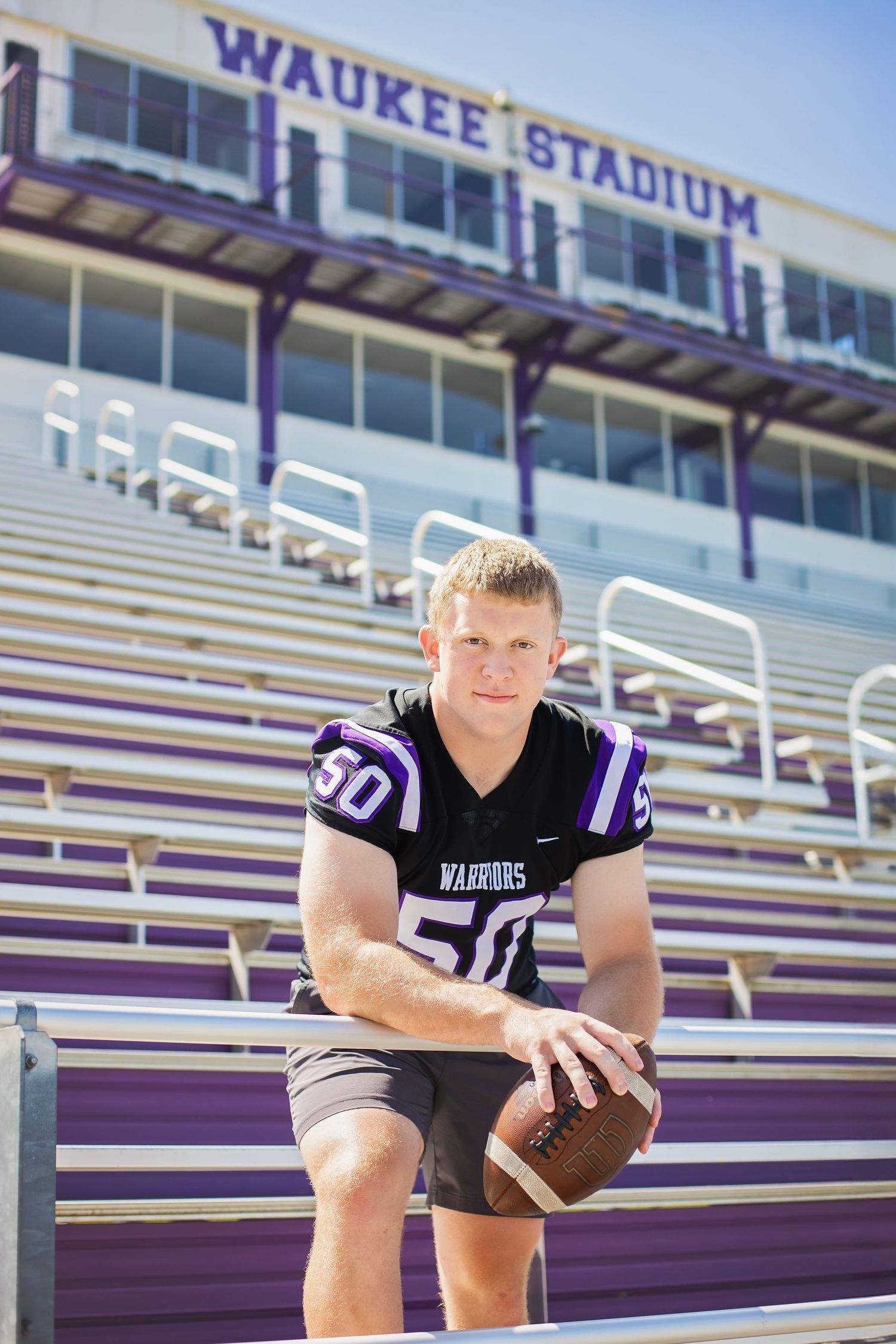 High school football player in Waukee Warriors jersey number 50 posing in stadium bleachers.