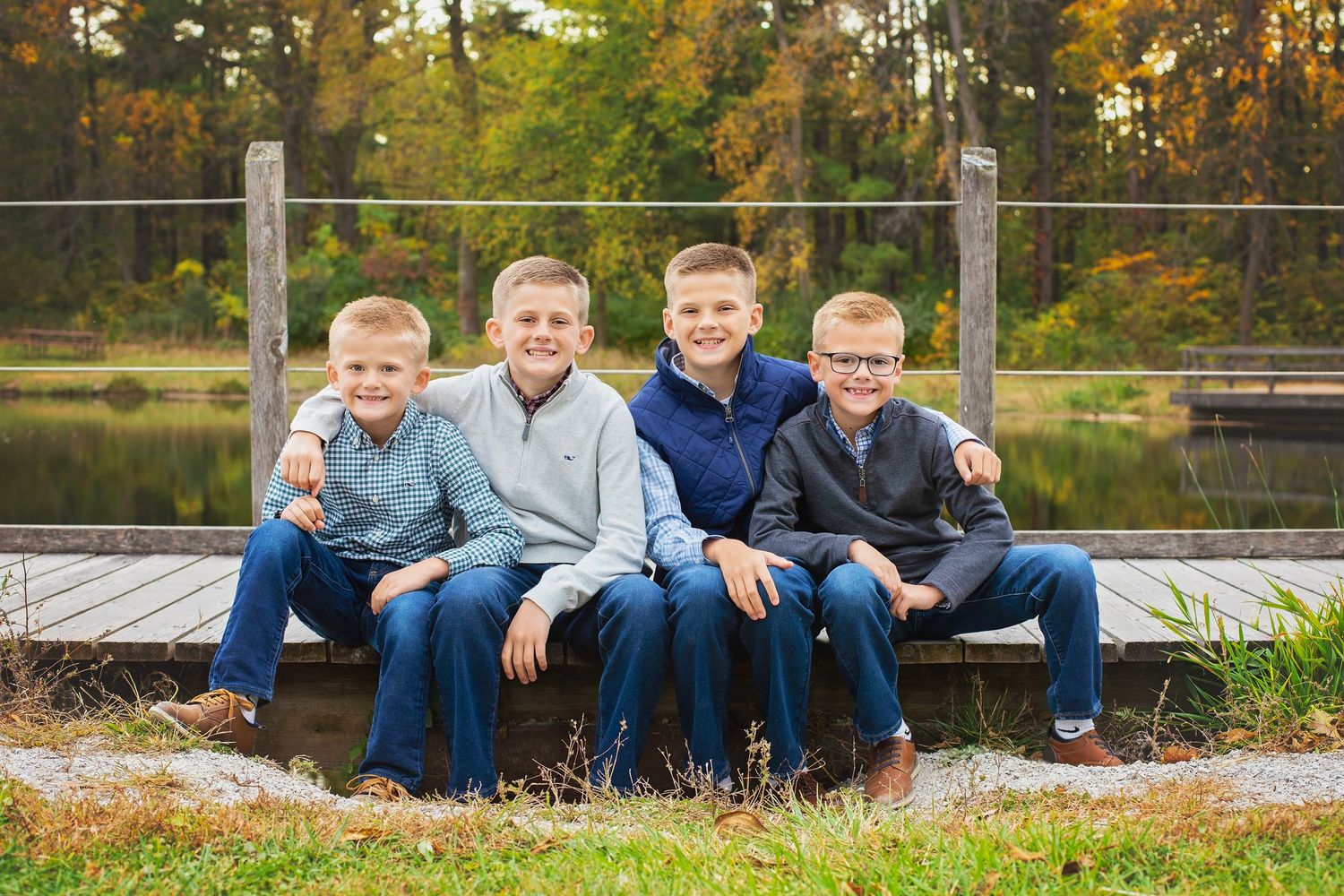 Four boys sitting on a wooden dock by a pond surrounded by autumn foliage.