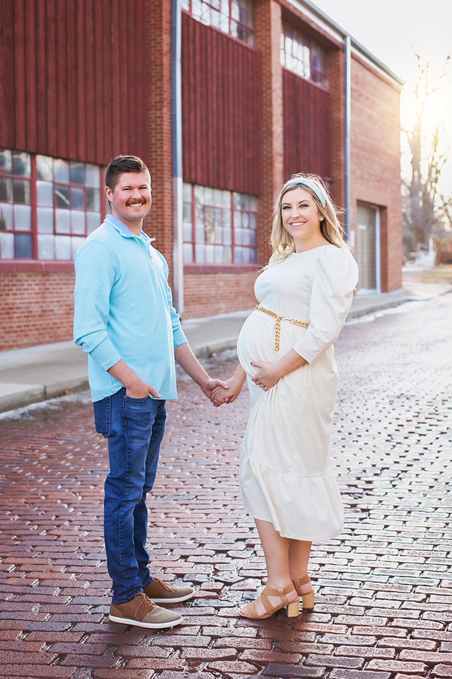 Pregnant couple holds hands on a cobblestone street, woman in white dress with gold belt, man in blue shirt.