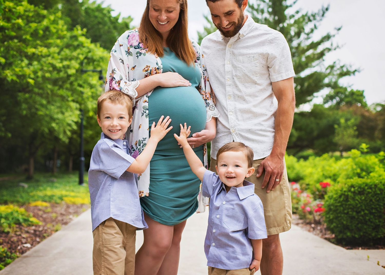 Pregnant woman in teal dress with husband and two young sons touching her baby bump.