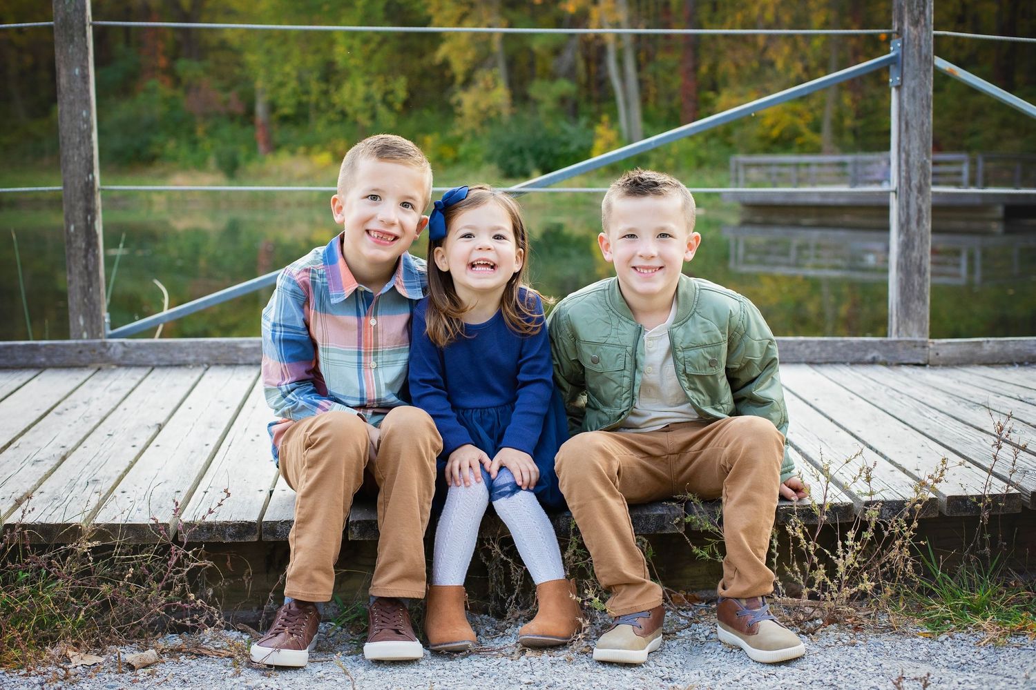 Three young siblings sitting and smiling on a wooden bridge outdoors.