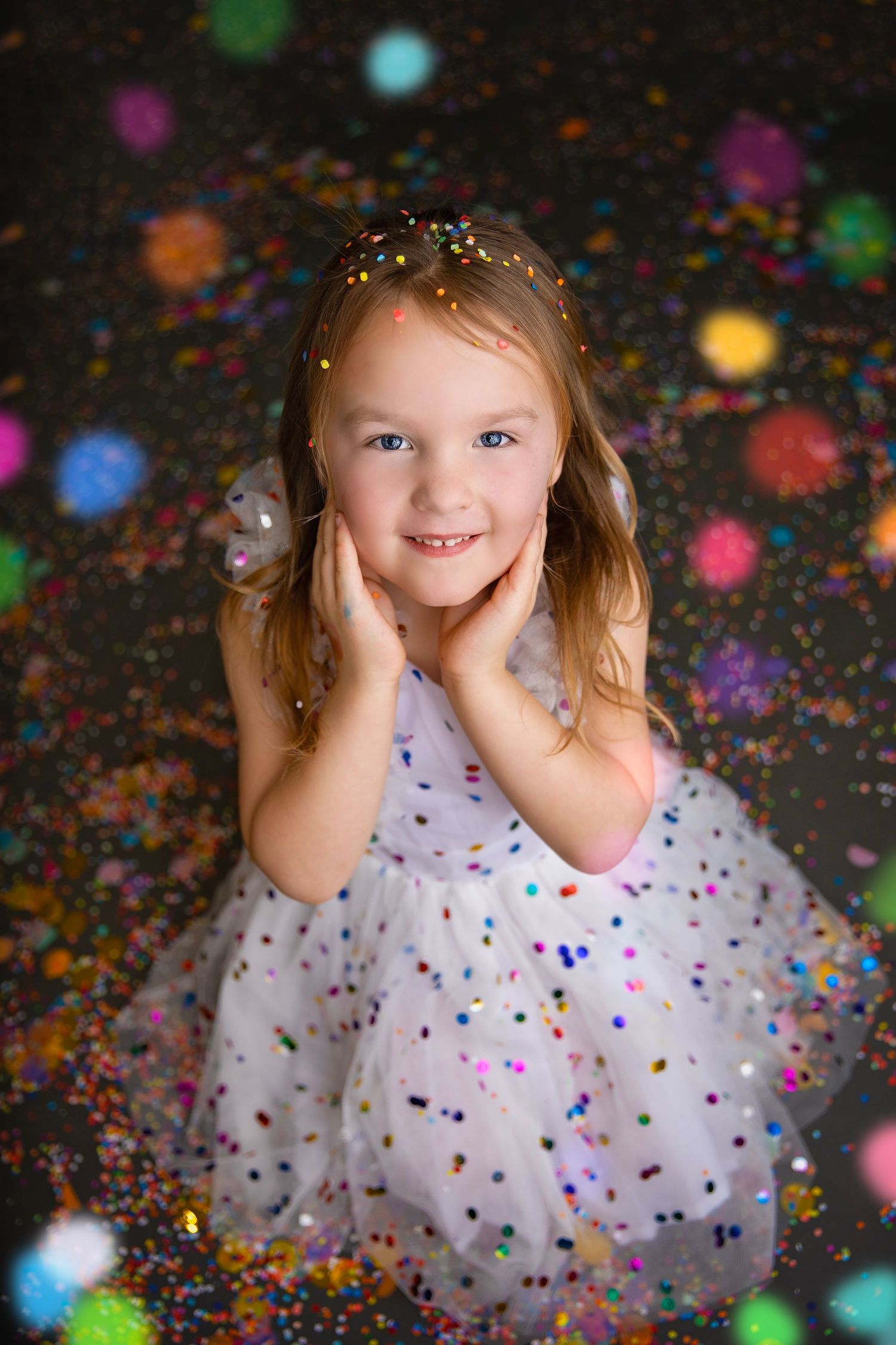 A young girl in a white polka dot dress poses with hands on cheeks surrounded by colorful confetti and bokeh lights.
