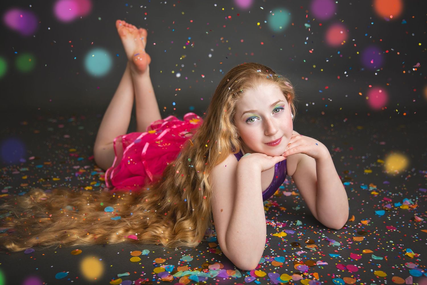 Young girl with long hair in pink and purple outfit lying on confetti-covered studio floor.